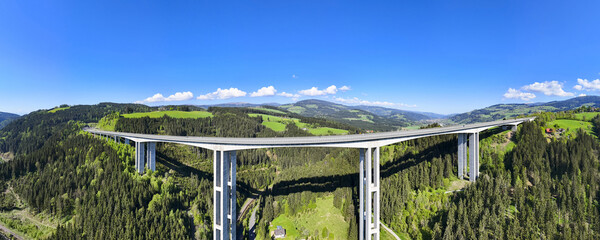 Aerial view of a soaring bridge, a concrete spine amidst the emerald hills and forests under a brilliant blue sky in Lavanttal, Wolfsberg, Carinthia.
