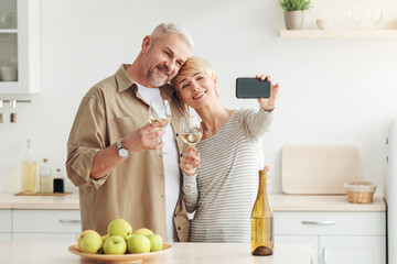 Selfie for holiday, app for phone, video call to family. Cheerful middle aged woman and man with wine on cozy kitchen interior, takes photo on smartphone with empty screen, free space