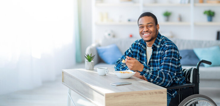 Cheerful black guy in wheelchair having breakfast, eating tasty cereal at home. Millennial with disability man enjoying healthy food in morning. Healthcare and good nutrition concept