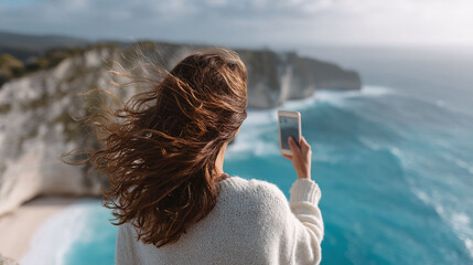 Woman captures stunning ocean view with her smartphone, hair blowing in the breeze. Serene travel, freedom and inspiration concept. Ideal for lifestyle or wanderlust themes.