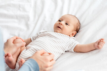 Baby Gymnastics. Unrecognizable Mother Making Straightening Exercises For Her Newborn Child At Home, Cute Infant Kid Lying On Bed, Looking At Mom And Smiling, Closeup Shot With Free Space
