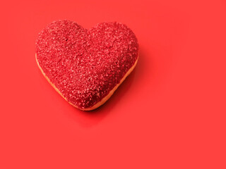 A heart-shaped doughnut with sprinkles on top sits on a red surface. The doughnut is the main focus of the image, and the red surface adds a touch of romance and sweetness to the scene