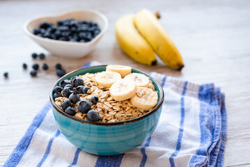 Porridge oatmeal with fresh fruit, a healthy breakfast bowl with oats, banana slices, and blueberries on a striped kitchen towel and rustic wooden table, with a spoon on the side.