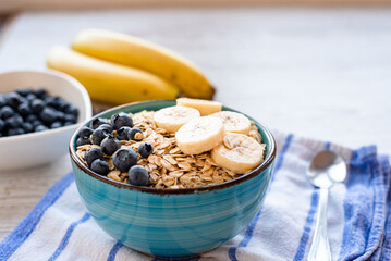 Porridge oatmeal with fresh fruit, a healthy breakfast bowl with oats, banana slices, and blueberries on a striped kitchen towel and rustic wooden table, with a spoon on the side.