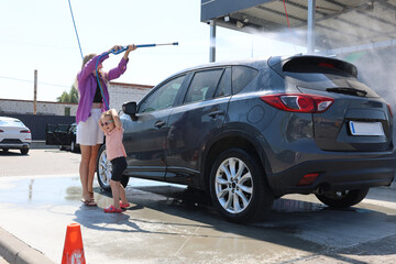 Child using a hose to clean a soapy car at an outdoor self-service wash station