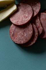 Pieces of sausage and a piece of cheese lie on a blue cutting board. The pieces of sausage are a rich red color with a marbled layer of fat. A rustic contrast to the modern look of the board.