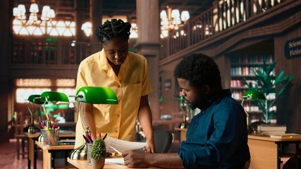 Black dedicated scholar works on their thesis project in the quiet bibliotheque, sharing ideas and comparing class notes for research completion. Achieving academic success. Camera A.