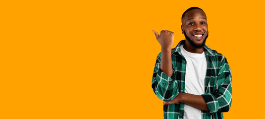 Look There. Cheerful Black Guy Pointing Thumb Finger Aside Advertising Your Text Standing Over Yellow Studio Background, Smiling To Camera. Advertisement Banner. Panorama With Copy Space