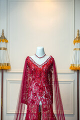 Elegant red bridal gown with floral lace and gemstone neckline, displayed on a mannequin in a traditional, formal Indonesian setting.