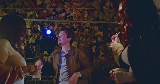 Friends enjoying a night party in the backyard. They are talking, laughing, and having drinks under the string lights.
