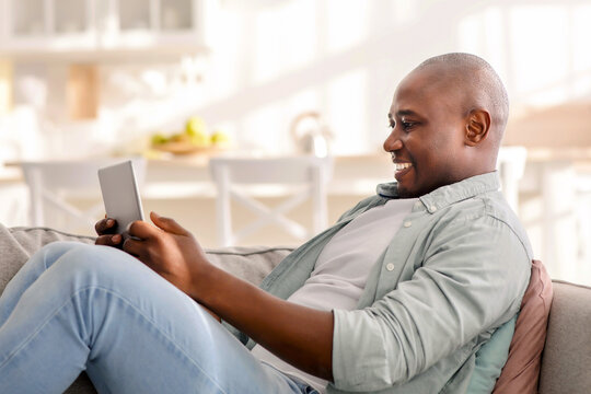 Happy mature african american man spending time with digital tablet at home, browsing internet, watching videos, checking social networks while relaxing on sofa in living room, copy space