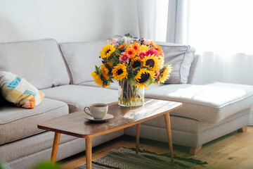 Cup of coffee and vase with huge multicolor summer flower bouquet on coffee table in modern cozy light living room with gray couch sofa. Open space home interior design. Cozy Design home decor.
