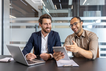 Two diverse businessmen collaborate in a modern office, reviewing a tablet with smiles, emphasizing teamwork and business success.