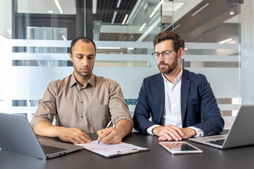 Two diverse men at a desk in an office, one signing a document while the other observes, laptops and tablet present.