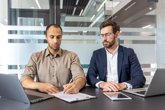 Two diverse men at a desk in an office, one signing a document while the other observes, laptops and tablet present. - Powered by Adobe