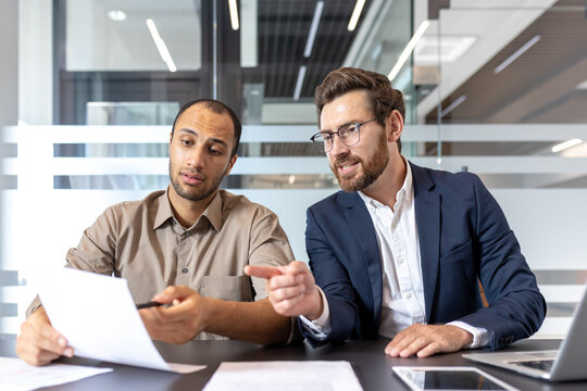 Two diverse businessmen collaborate, reviewing documents at a modern office desk, discussing and analyzing data for a project.