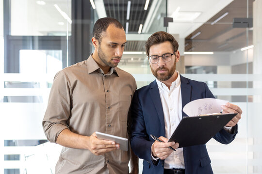 Two business professionals, one holding a tablet, the other examining a document, engaged in a discussion within a modern office setting. - Powered by Adobe