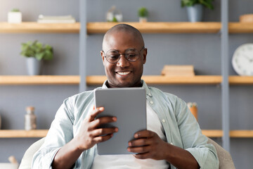 Happy black mature man using digital tablet, sitting in armchair over shelves with accessories, reading book online or surfing internet on pad at home. Gadgets lifestyle, technology concept