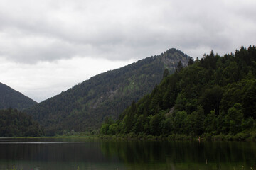 Serene mountain lake under a dramatic overcast sky, surrounded by lush green forested hills. The calm water reflects the surrounding nature, creating a peaceful and moody atmosphere 