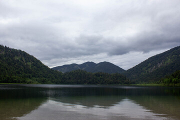 Serene mountain lake under a dramatic overcast sky, surrounded by lush green forested hills. The calm water reflects the surrounding nature, creating a peaceful and moody atmosphere 