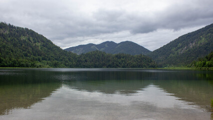 Serene mountain lake under a dramatic overcast sky, surrounded by lush green forested hills. The calm water reflects the surrounding nature, creating a peaceful and moody atmosphere 