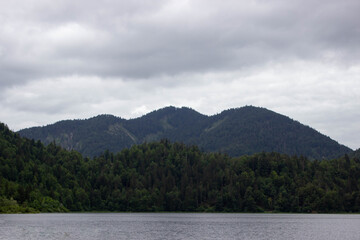 Dark green forested mountains surround a peaceful alpine lake under an overcast sky. The calm water reflects the landscape, evoking a quiet, moody, and untouched natural atmosphere.