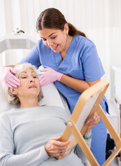 Fototapeta premium Young female dermatologist showing elderly female client result of facial beauty procedure in mirror in esthetic medicine center