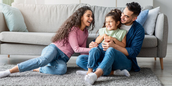 Middle Eastern Parents Having Fun With Their Little Daughter At Home, Arabic Mom And Dad Tickling Kid And Laughing Together In Living Room, Enjoying Domestic Leisure, Copy Space, Panorama - Powered by Adobe