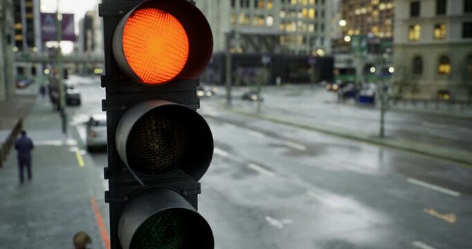 A bright orange traffic light glows in the foreground while cars navigate through slick streets. Raindrops create reflections on the ground, adding texture to the urban landscape at dusk.