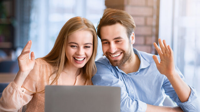 Young couple communicating online on laptop at cafe, waving to camera. Millennial woman and her boyfriend greeting family or friends, talking to someone on webcam at restaurant