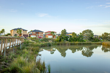 Obraz premium A scenic and peaceful recreational space of Tarneit Lakes in Melbourne, Australia, with calm water reflecting nearby modern houses and a wooden boardwalk or walking trail alongside.