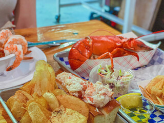 A colorful seafood platter with a whole lobster, shrimp cocktail, lobster roll, chips, and coleslaw on a rustic wooden table.