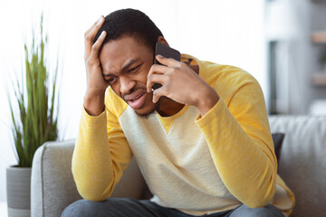 Closeup of sad black guy sitting on couch, having conversation on mobile phone, touching his head. Upset african american young man talking on smartphone, fighting with girlfriend