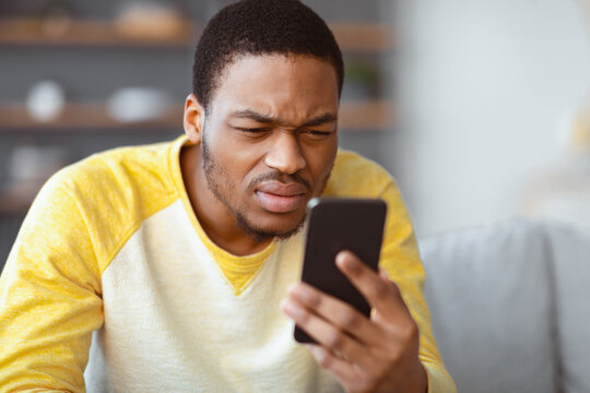African american guy squinting while looking at mobile phone screen at home, copy space, blurred background. Young black man reading suspicious message on his smartphone, closeup