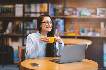 Carefree latin woman working distantly on laptop computer, sitting at cafe and enjoying coffee, free space. Positive lady holding mug with hot beverage, looking aside and smiling