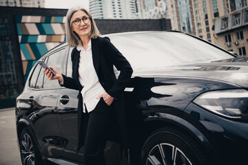 Successful mature businesswoman with grey hair and elegant outfit standing next to a car in an urban street setting