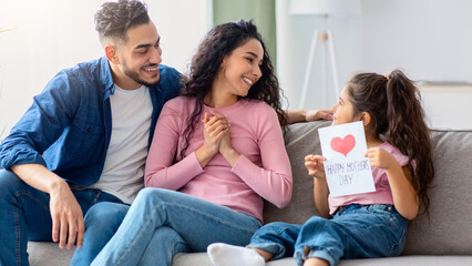 Happy Mother's Day Concept. Smiling little girl holding handmade greeting card with drawn red heart for her mom, congratulating her mommy while they sitting together on couch in living room