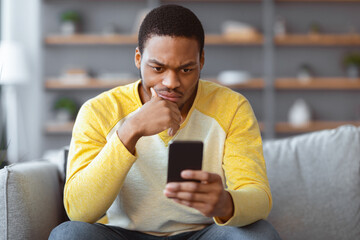 Closeup of thoughtful black guy sitting on sofa at home, using smartphone, checking new...