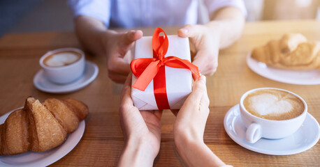 Young couple with gift at coffee shop, having lunch together, celebrating anniversary, closeup of hands. Millennial boyfriend giving birthday present to his beloved woman at cafe, cropped view