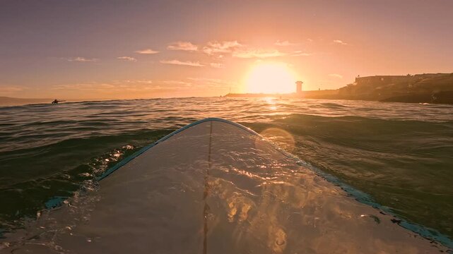POV, FPV, LENS FLARE: Surfer on surfboard waiting in lineup for riding a wave. Setting sun creates a golden reflection on the surface of wavy water during an evening surf along the Moroccan coast.