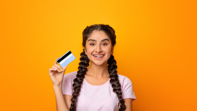 Easy Digital Cashless Payment. Portrait of smiling casual young asian woman holding credit card in hand and looking at camera, thinking about sales and purchases, posing over yellow studio background