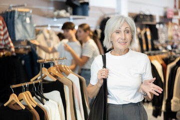 Mature woman buyer noticed desired product in clothes store and happily smiles from upcoming purchase. Elderly female buyer looks around in showroom and chooses right fit outfit.