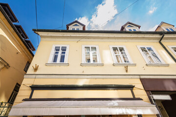 Low angle view of a pastel yellow building facade in Ljubljana old town, Slovenia, featuring white framed windows, dormer gables and shop awning under a blue summer sky with scattered clouds.