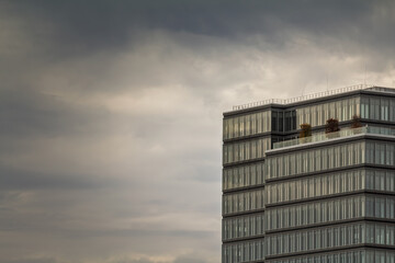 Glass and steel office block with repetitive windows rises against a moody, overcast sky. The minimalist facade suggests corporate architecture and urban density under changing weather.