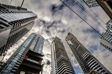 Upward view of condo towers under construction in downtown Toronto, Ontario. Cranes, glass facades and overhead wires frame a dense skyline, in an area of Canada being redeveloped for real estate.