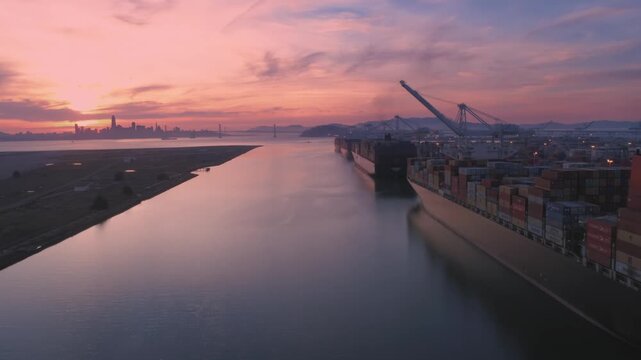 Cargo ships docked at the Port of Oakland, are being loaded with shipping containers at sunset. The city skyline is visible in the distance. Oakland, California, USA. 12 March 2025