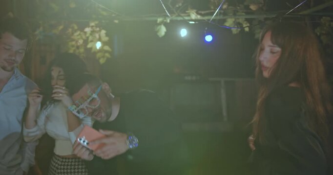 Friends gather in the backyard at night, enjoying drinks, music, and each other's company under string lights.