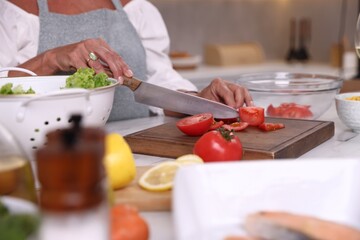 Senior woman cutting tomato at table in kitchen, closeup