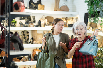 Mature woman with young daughter in store eye windowshopping product she likes and tactilely checks authenticity and naturalness of leather material from which bag is made.