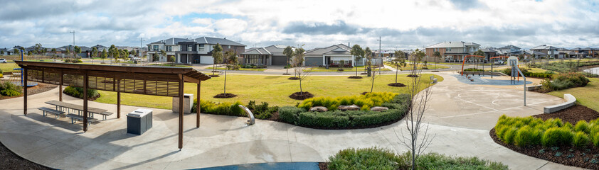 Panoramic view of a modern suburban park in a new residential estate in Melbourne, Australia, with a sheltered picnic area, open lawn, and a basketball half-court, and residential houses nearby. © Doublelee